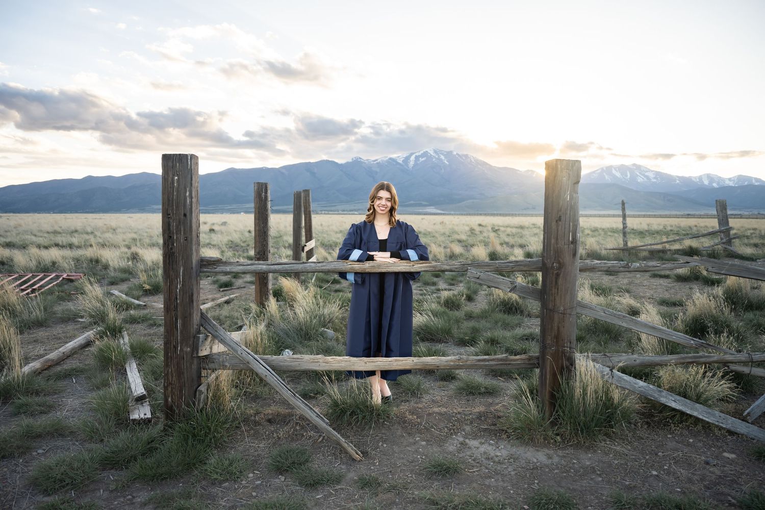 High school senior girl standing between rustic wooden fences with mountain views during a golden hour senior photography session in Utah.