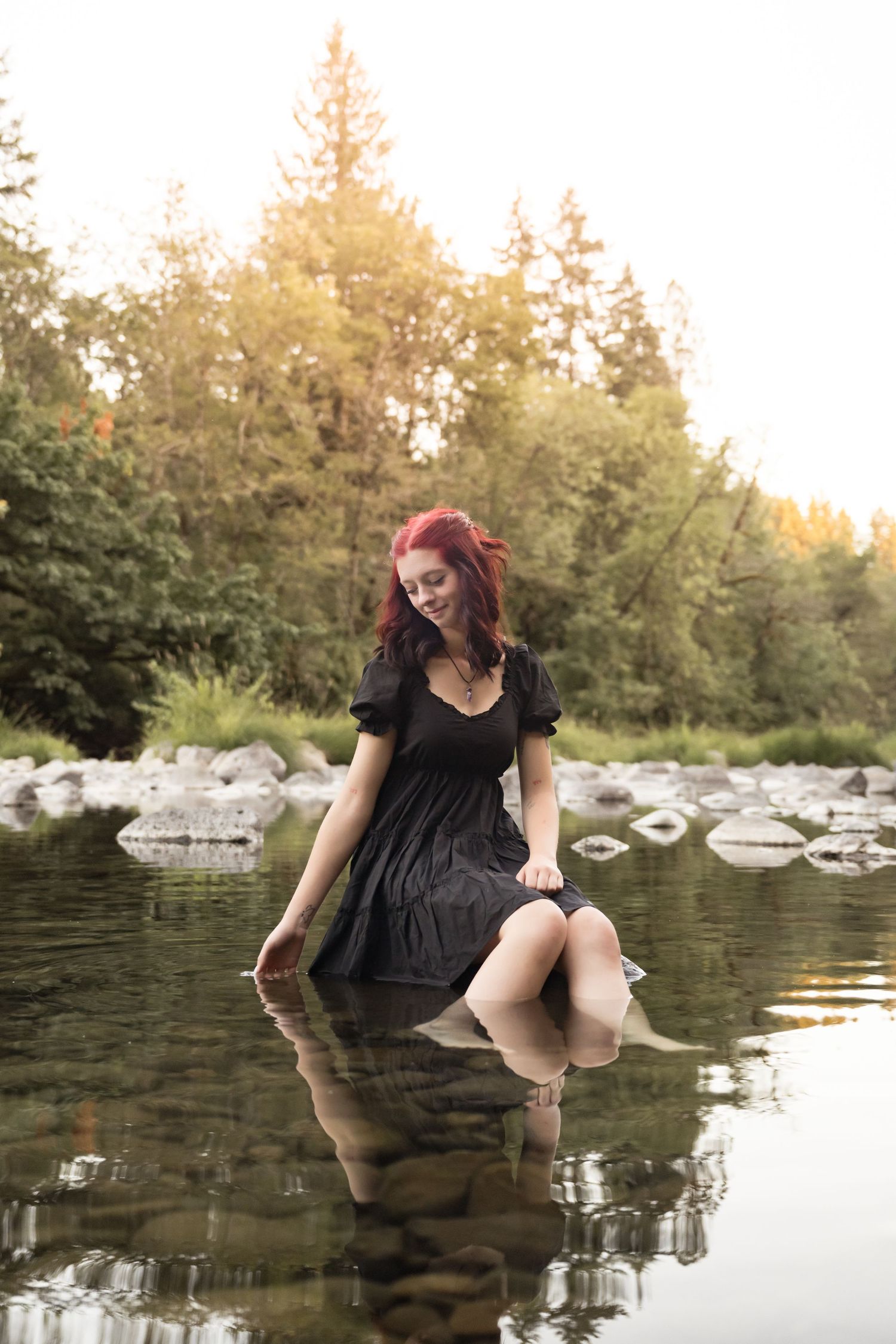 High school senior girl standing in a calm river surrounded by evergreen trees during an outdoor senior photography session in Washington