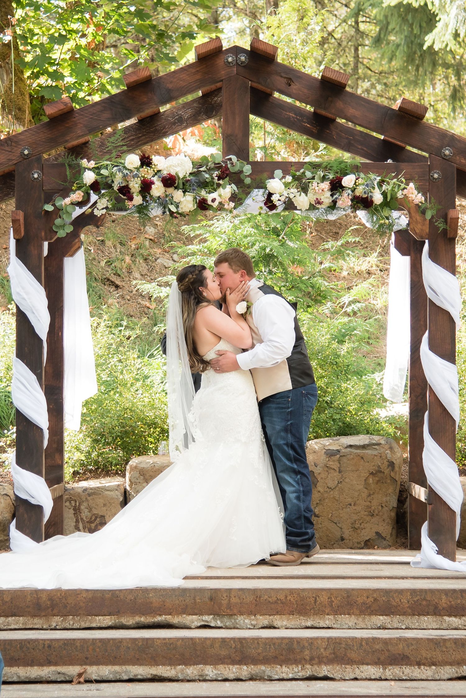 Newly married couple embracing beneath a wooden wedding arbor in a scenic outdoor ceremony.