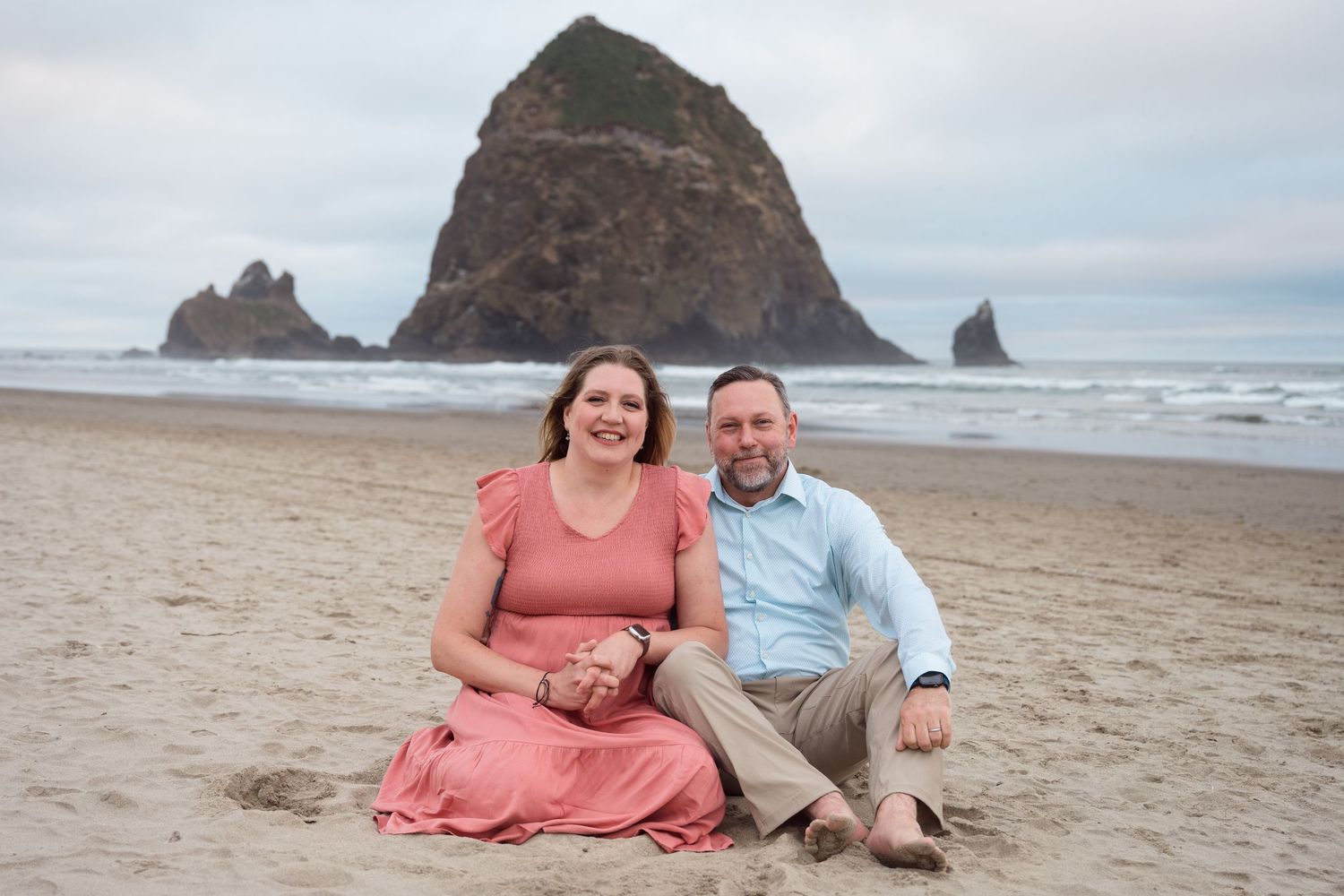 Couple sitting together on the Oregon Coast with Haystack Rock during a romantic beach portrait session.