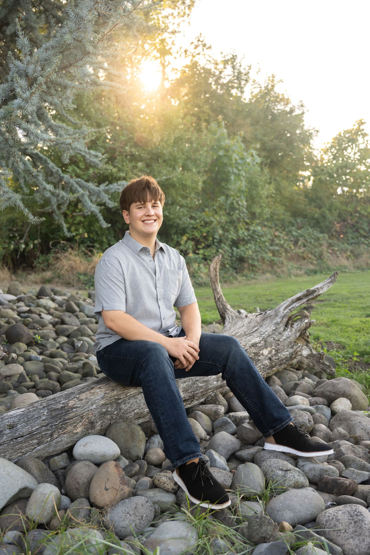 High school senior boy sitting on river rocks surrounded by trees during a relaxed outdoor senior portrait session in Washington.