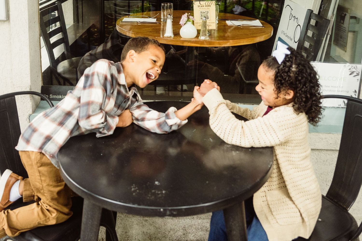 Twin siblings sharing a playful moment at an outdoor bistro table during an lifestyle family photography session.
