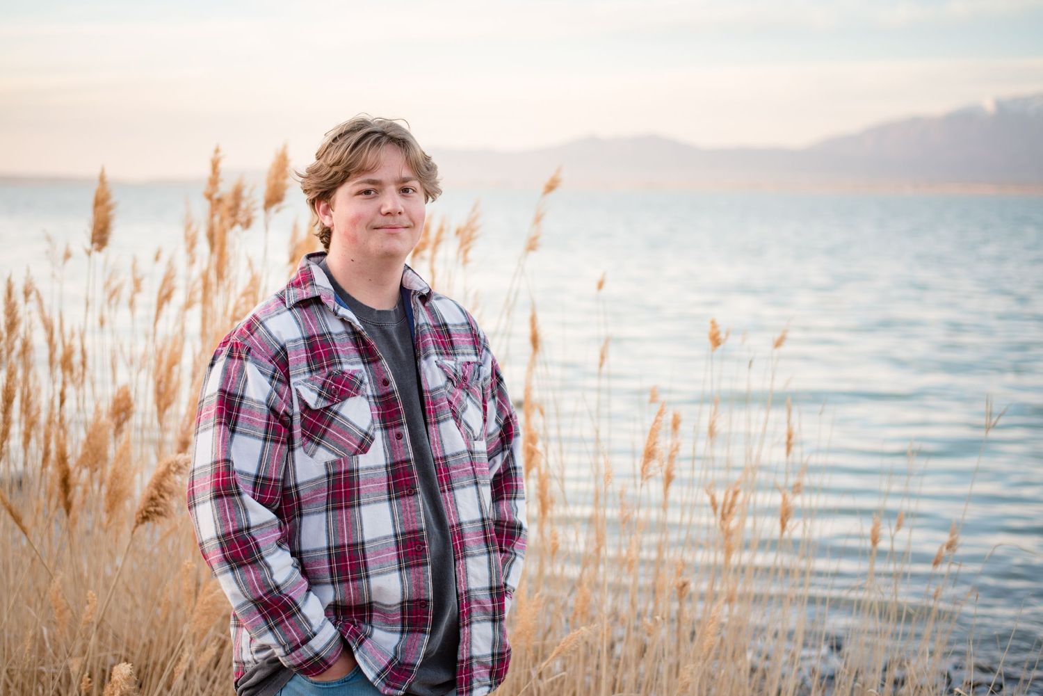 High school senior boy standing beside a lakeshore with tall reeds during a natural light outdoor senior photography session in Utah.