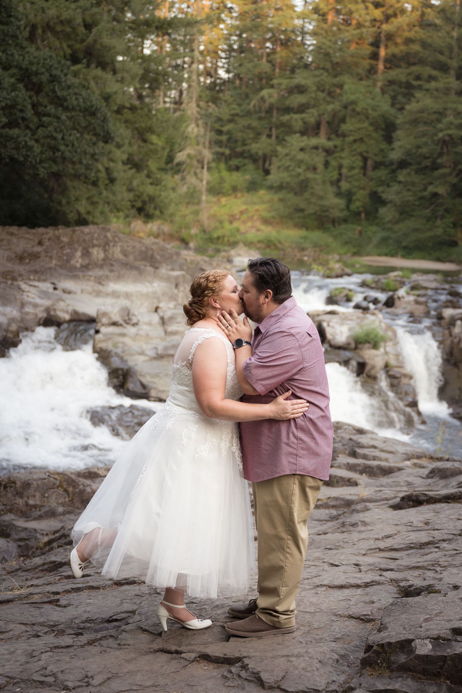 Bride and groom embracing beside a scenic waterfall during a romantic outdoor wedding portrait session.