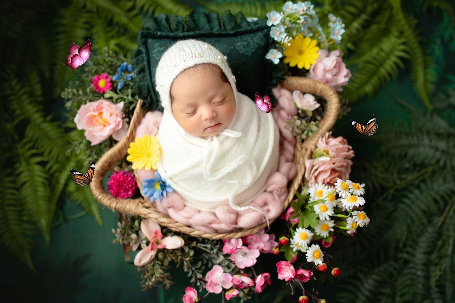 Newborn sleeps peacefully in basket surrounded by colorful spring flowers and green ferns in overhead photo.