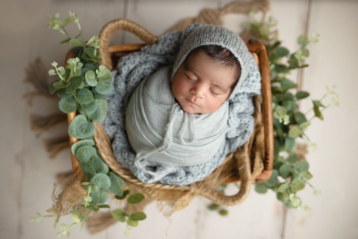 Newborn baby sleeping peacefully in wooden basket surrounded by delicate green eucalyptus leaves and soft blankets.