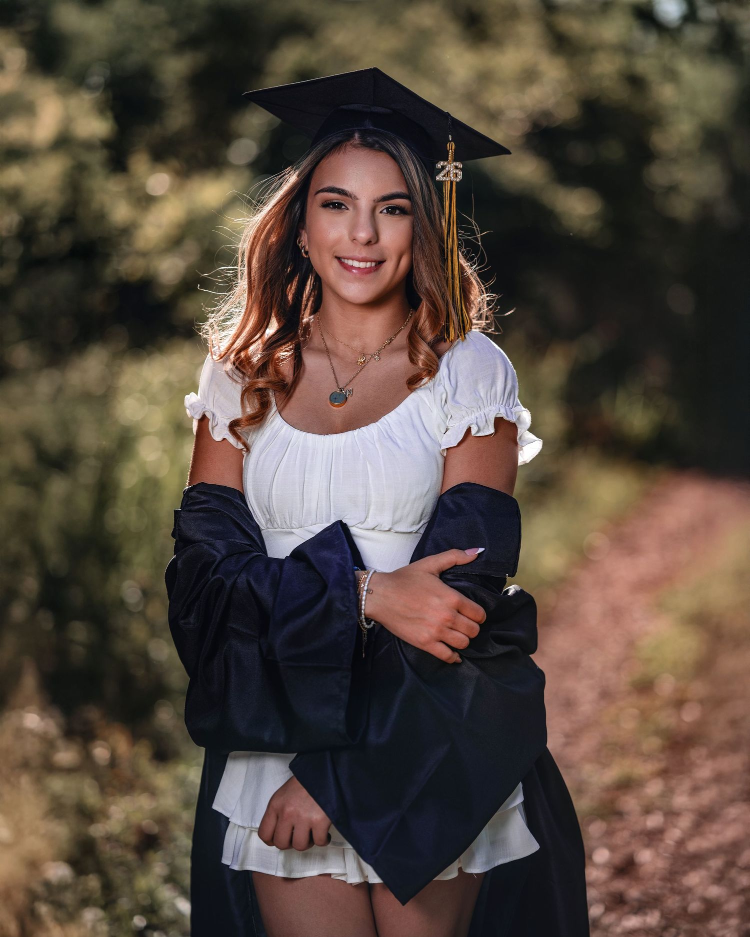 Smiling graduate in white dress and navy cap and gown poses outdoors on a sunlit path.
