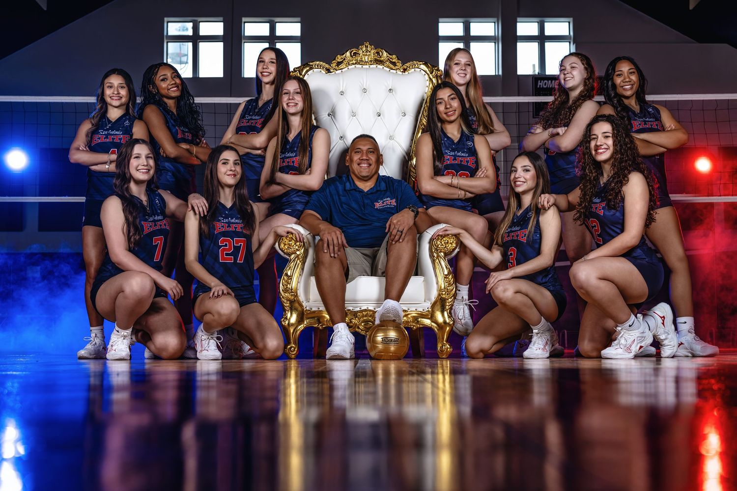 Women's basketball team poses with coach on gold throne in navy uniforms on indoor court with dramatic blue and red lighting.