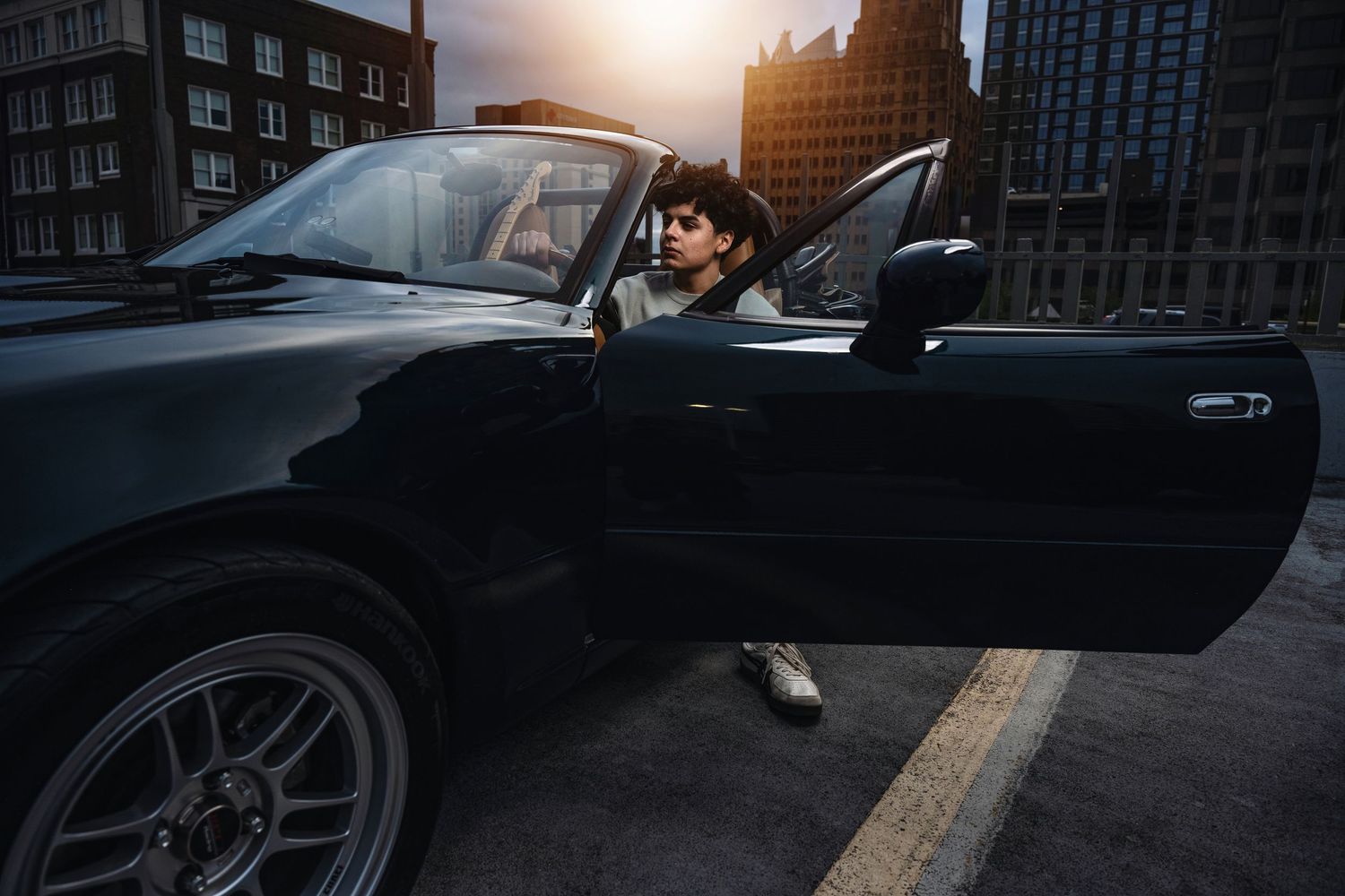 Man in suit sitting in black convertible car with city buildings in background at sunset.