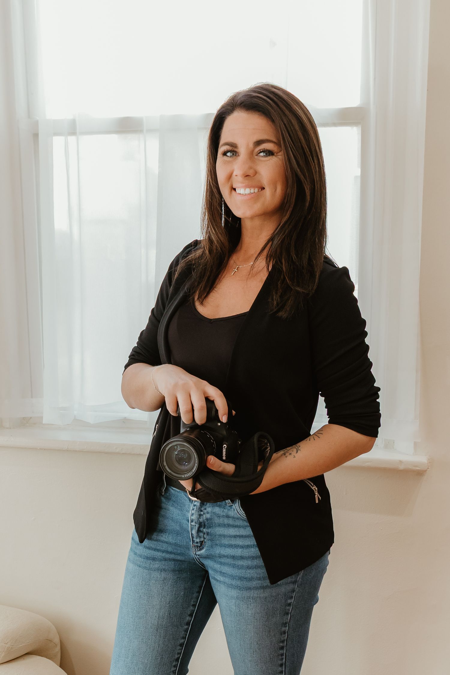 Woman in black top and blue jeans poses casually against white wall in bright interior setting.
