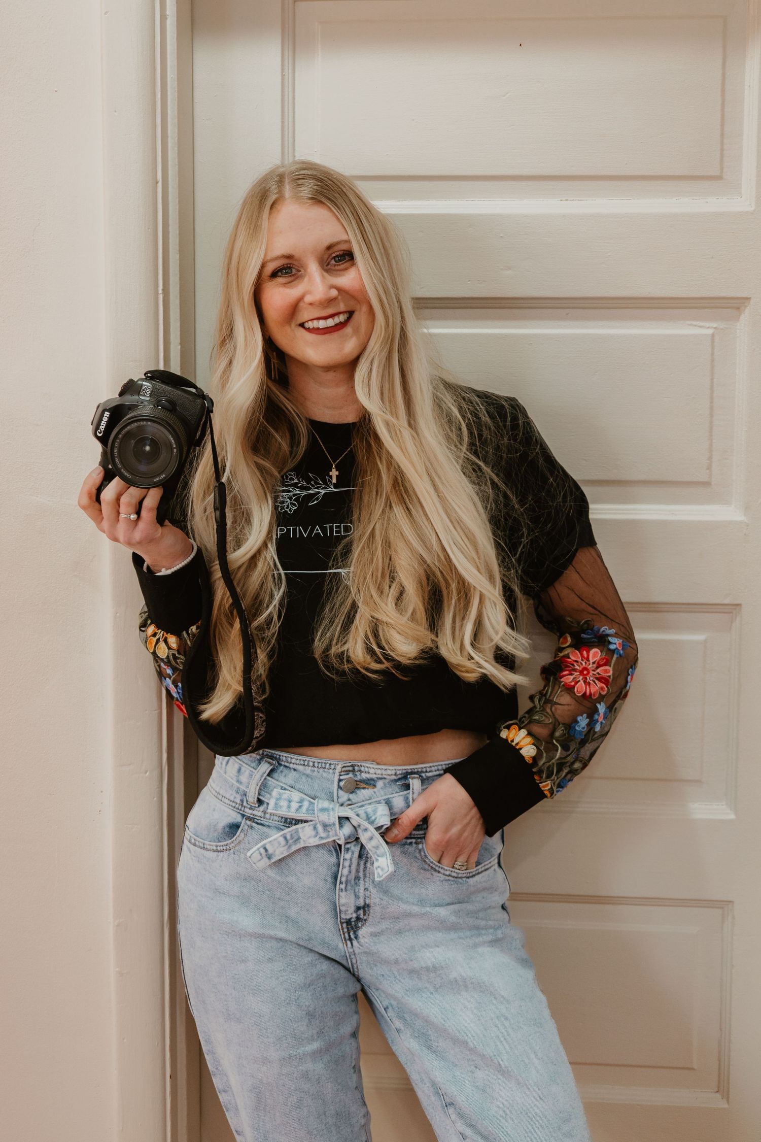 A photographer in a floral embroidered jacket and light wash jeans holds a camera while posing against a white door.