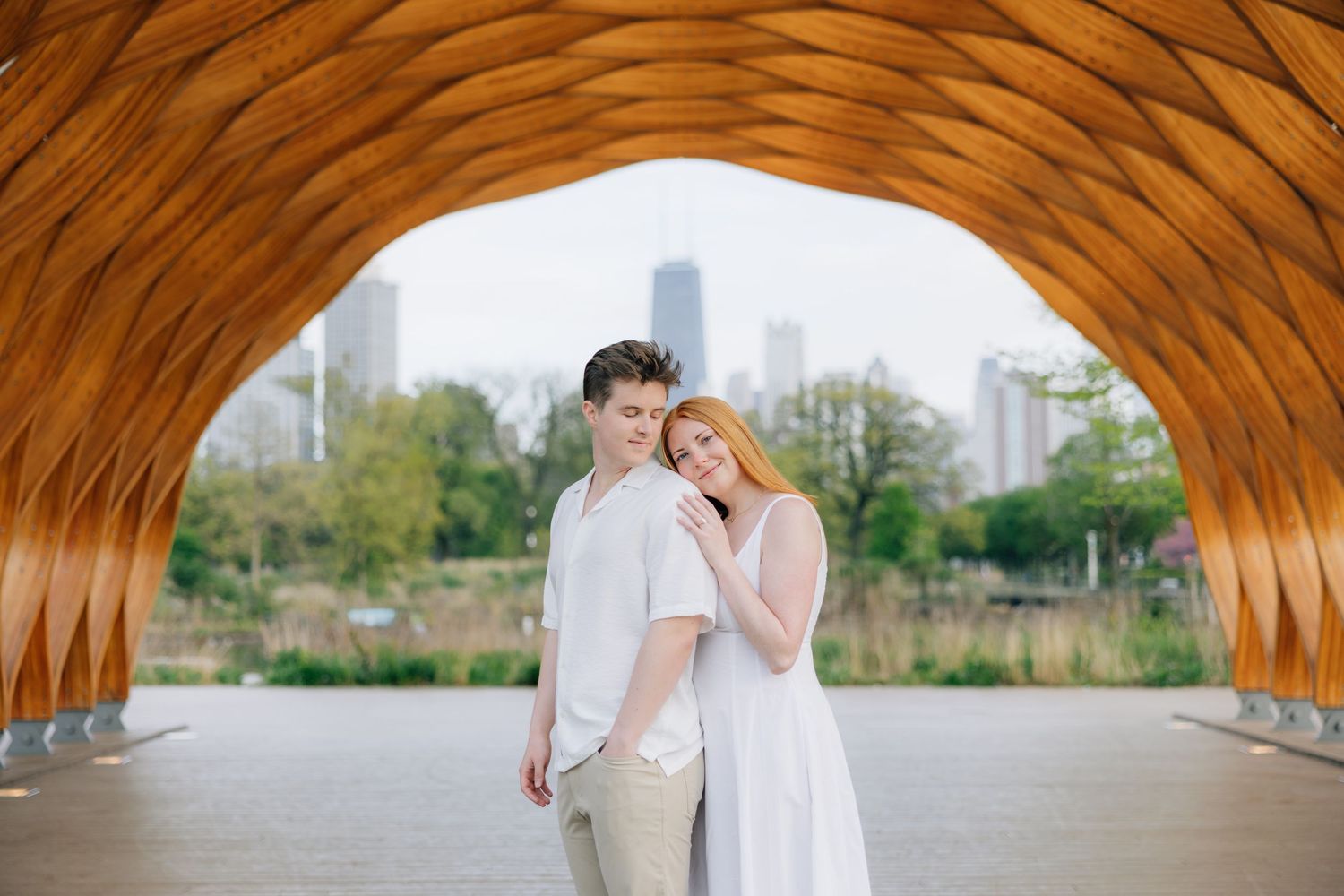 Couple embracing under wooden arch structure with Chicago skyline in background.