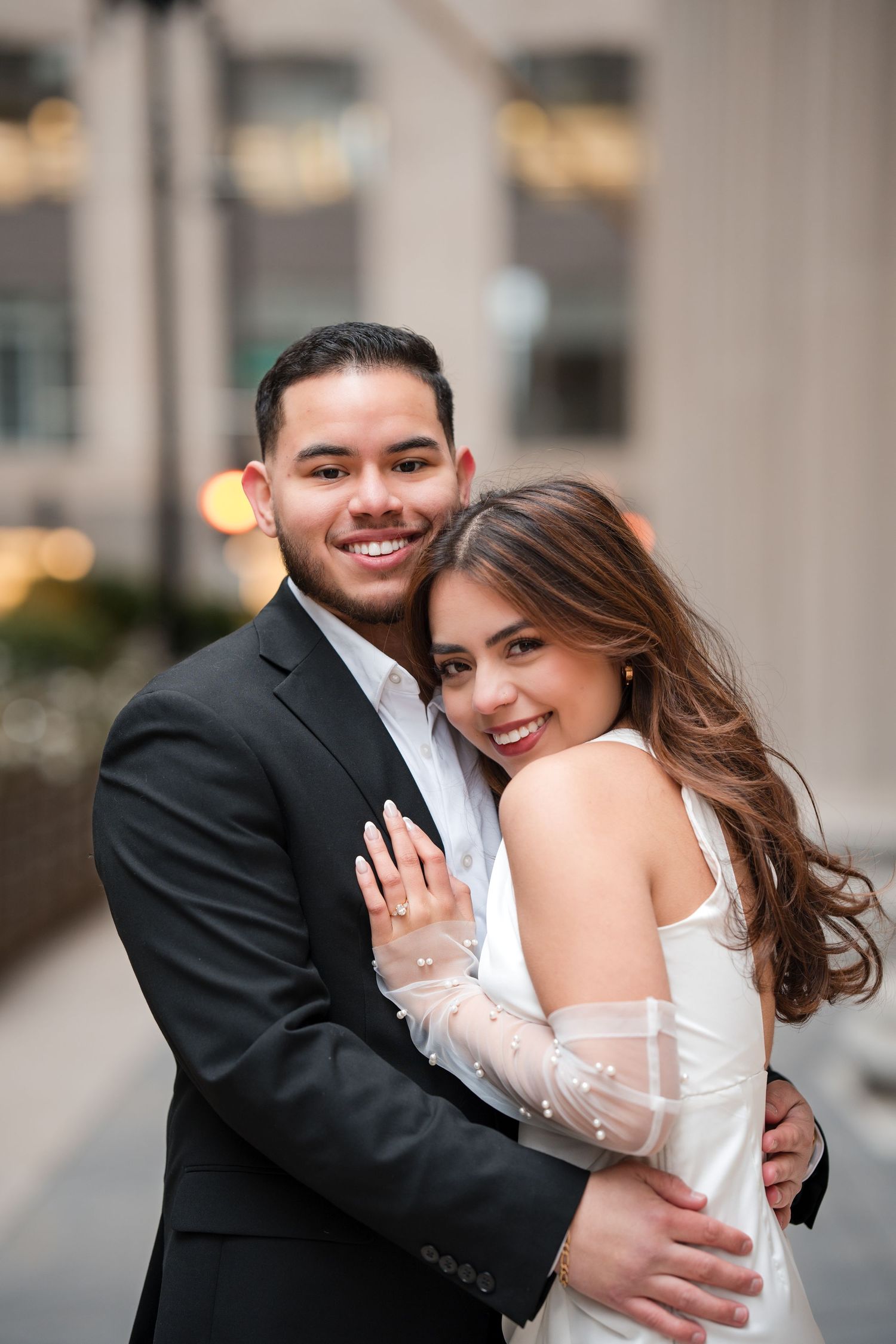 A smiling couple in formal attire embraces outdoors, the woman in a white gown and the man in a black suit.