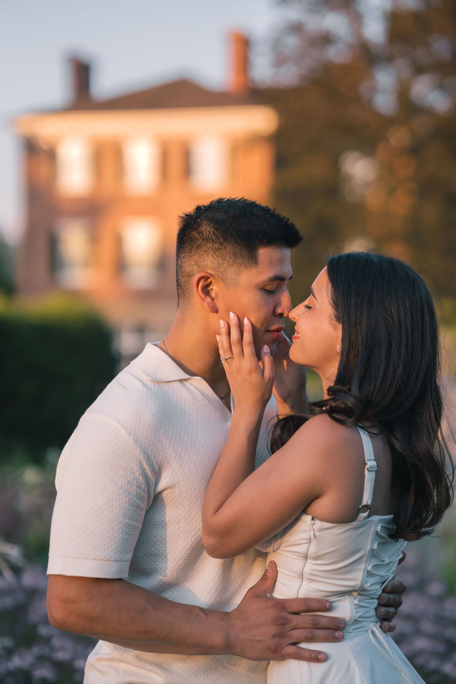Couple sharing a romantic moment at golden hour with a blurred brick mansion in the background.