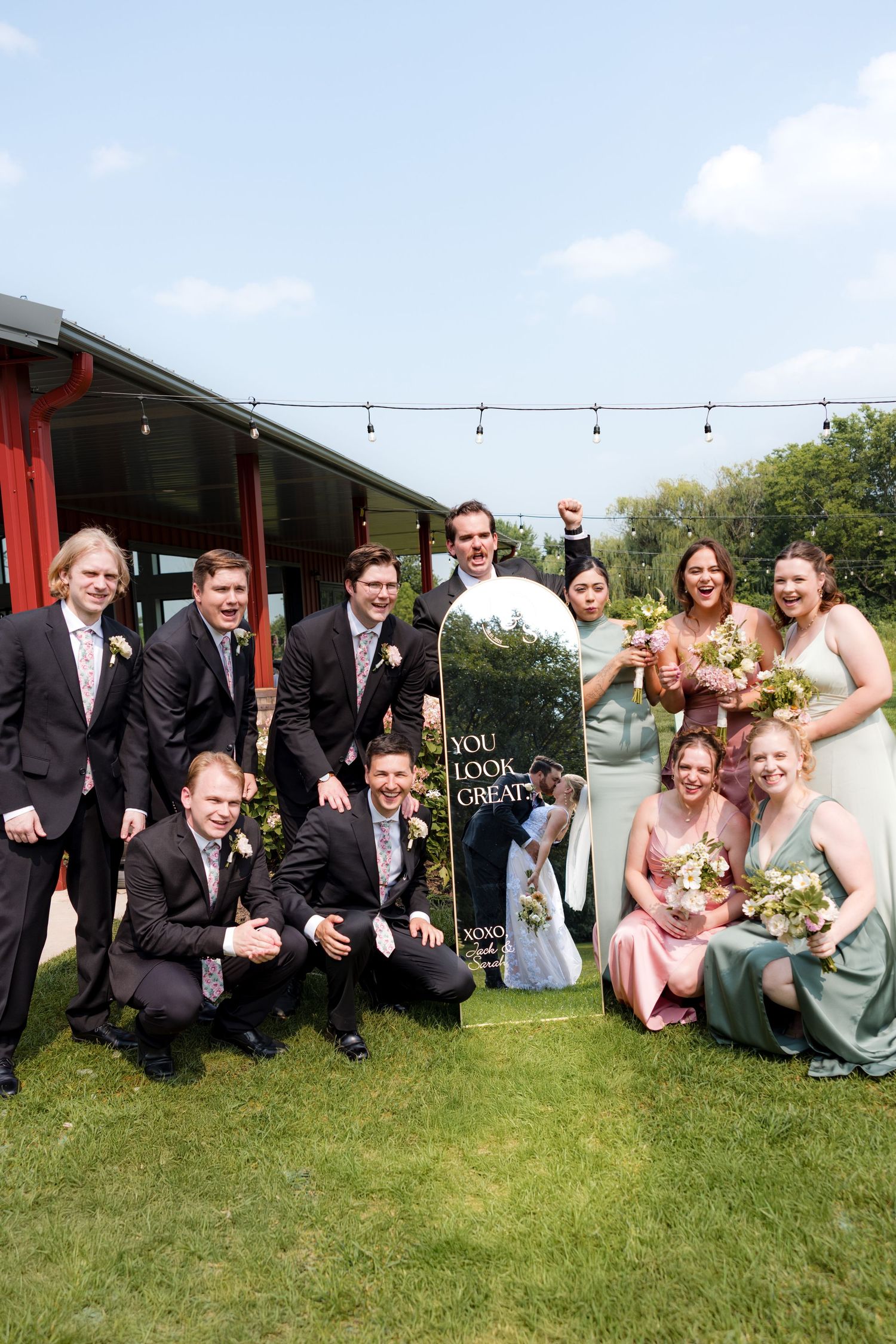 Wedding party poses together on grass with red barn in background during outdoor celebration.