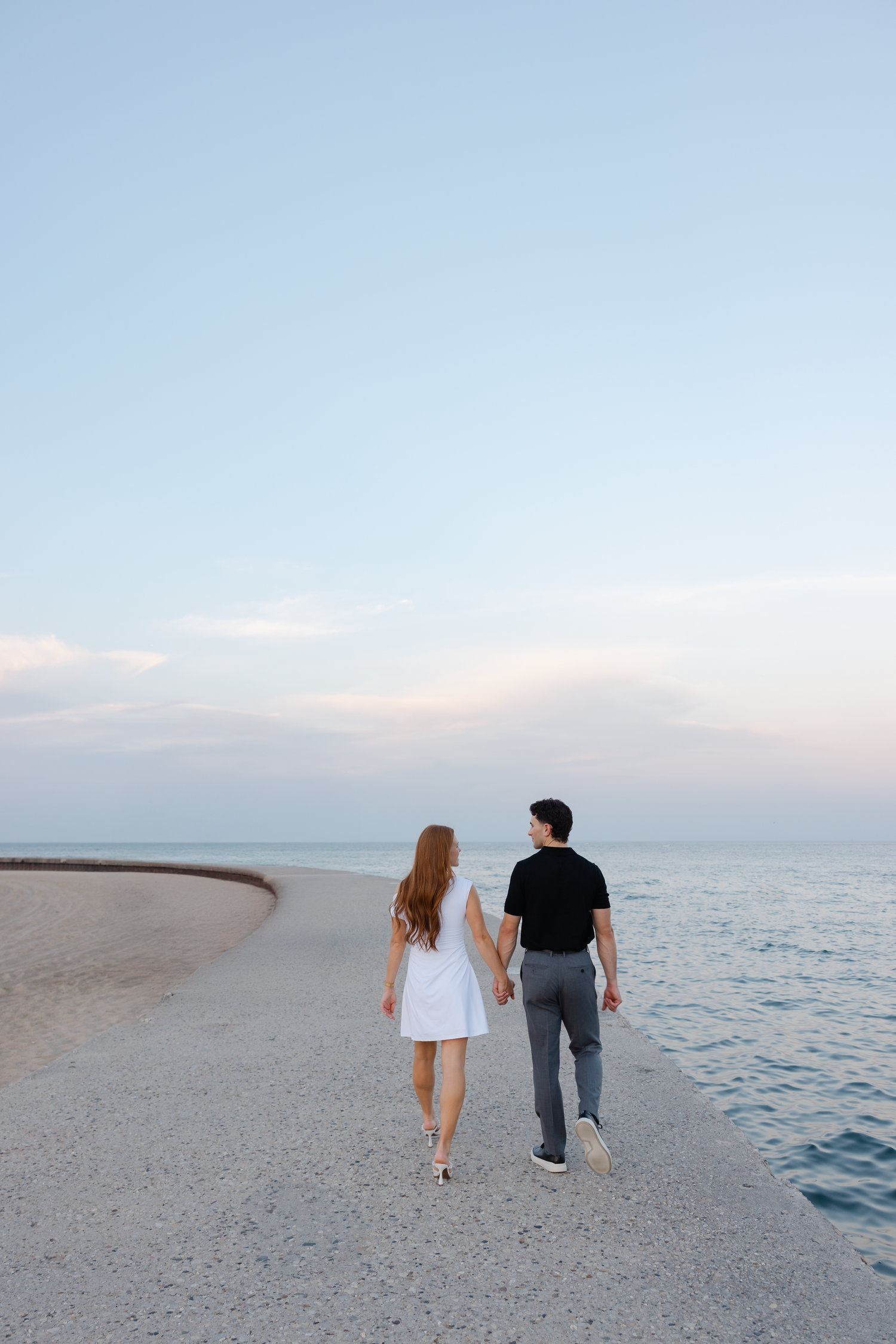 Couple holding hands walking along a sandy beach shoreline at dusk with calm ocean waters.