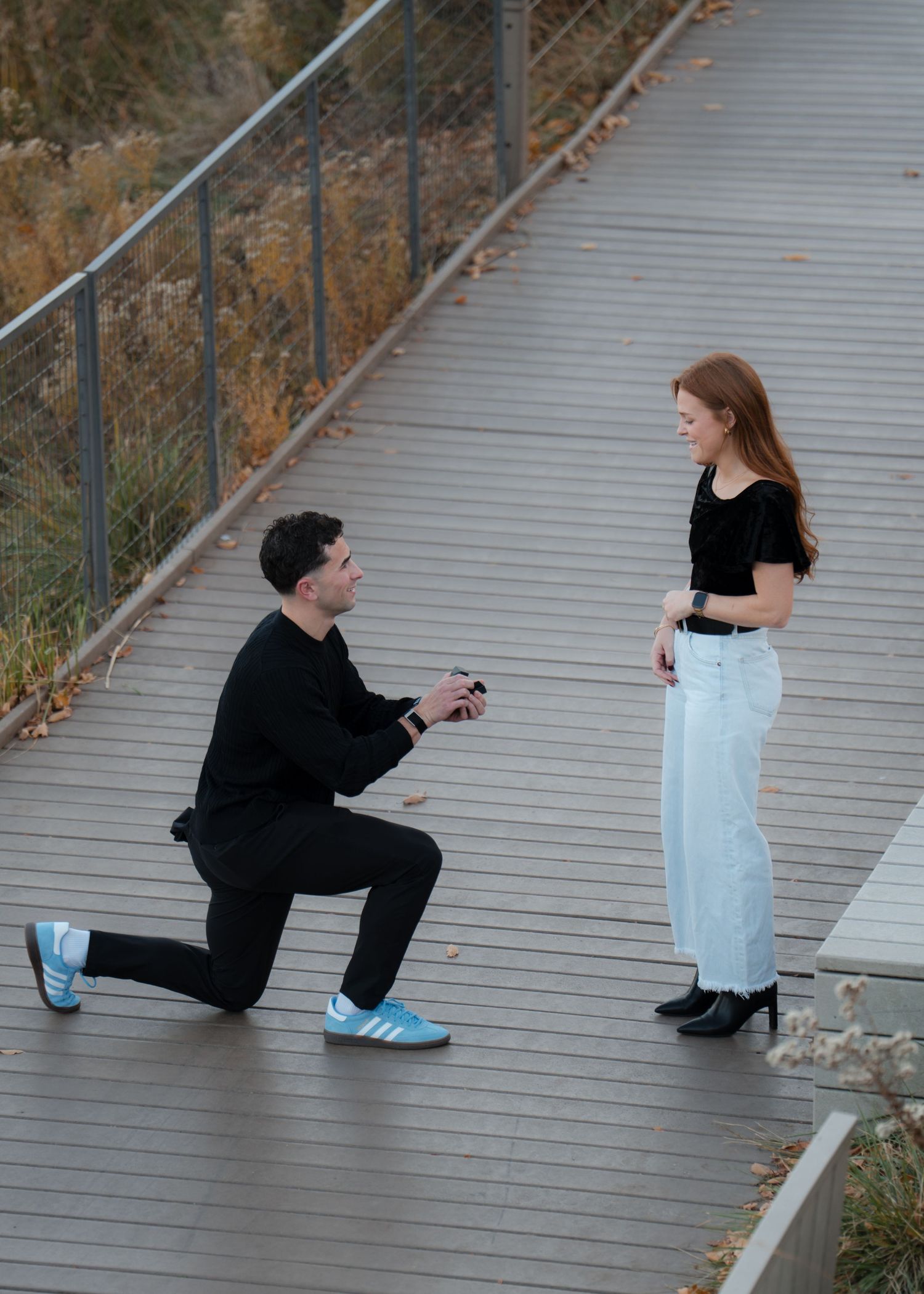 Man kneeling on one knee proposing to a woman on a wooden bridge outdoors in autumn.
