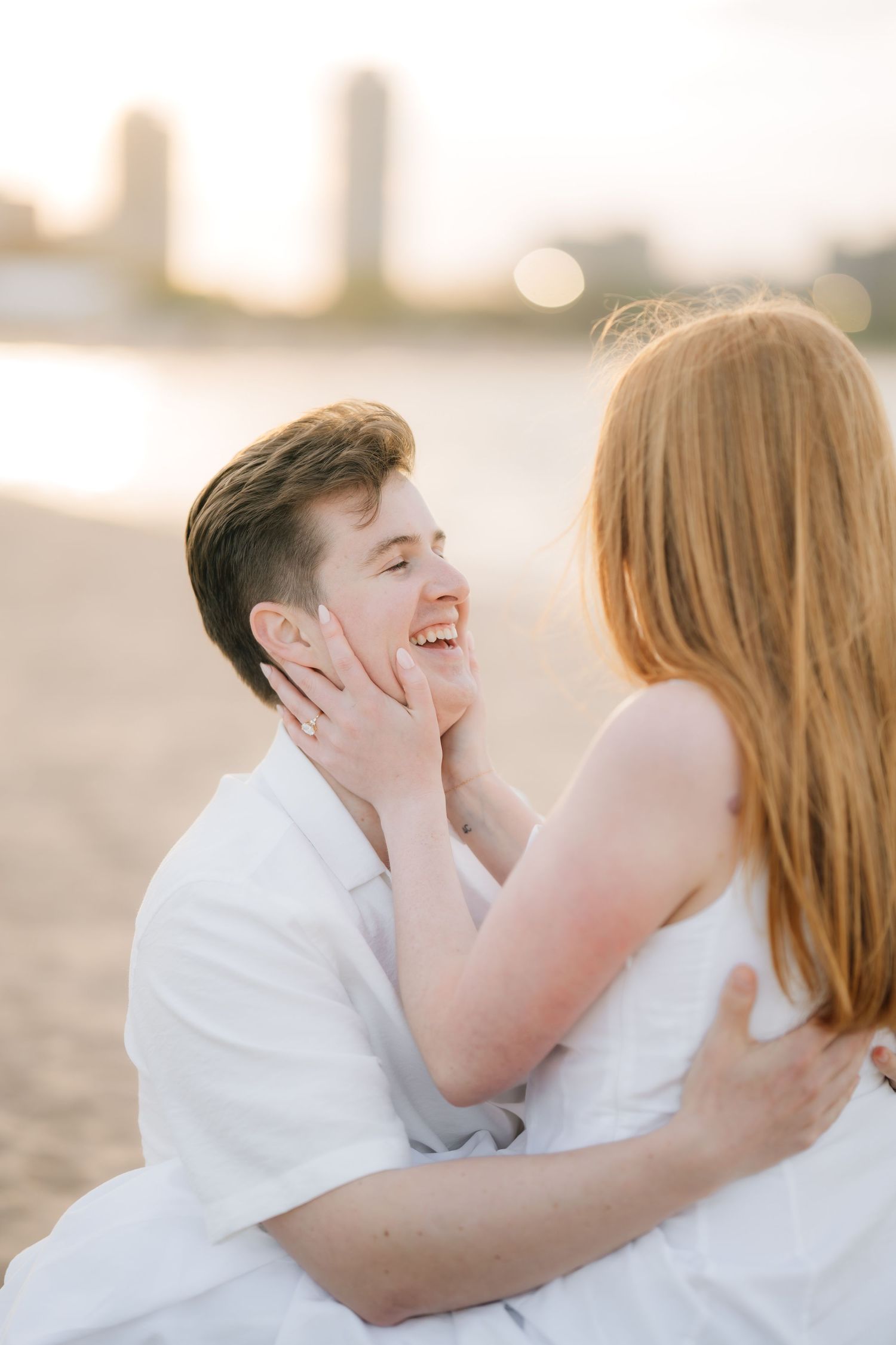 Couple sharing a tender moment on the beach at golden hour, woman touching man's face as he laughs.