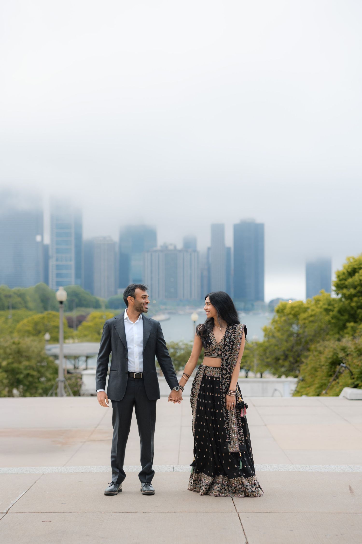 Indian couple in traditional and formal attire holding hands with misty city skyline backdrop.