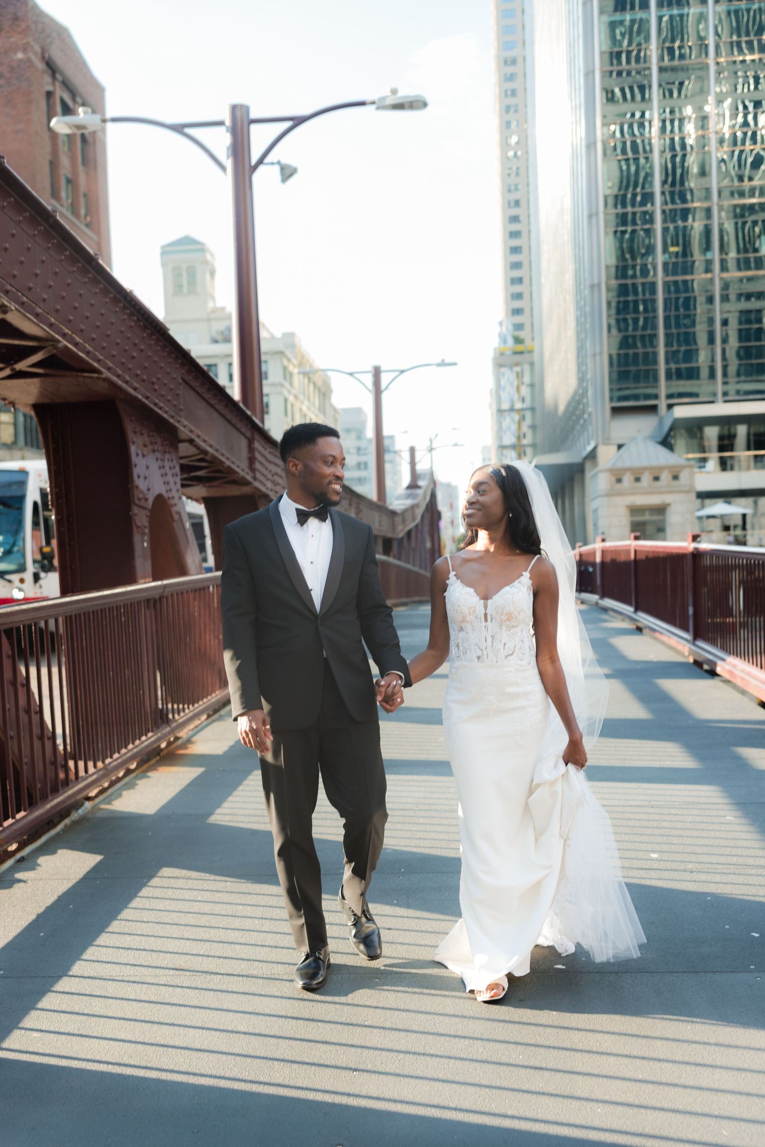Newlyweds walk hand in hand across urban bridge with city architecture in background.