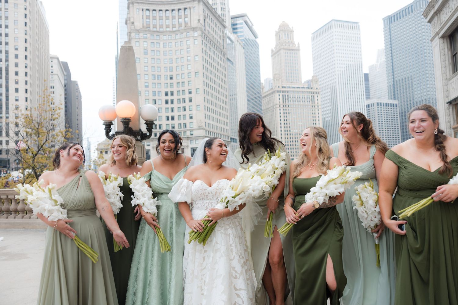 Wedding party in sage green dresses and formal attire poses together against urban backdrop.