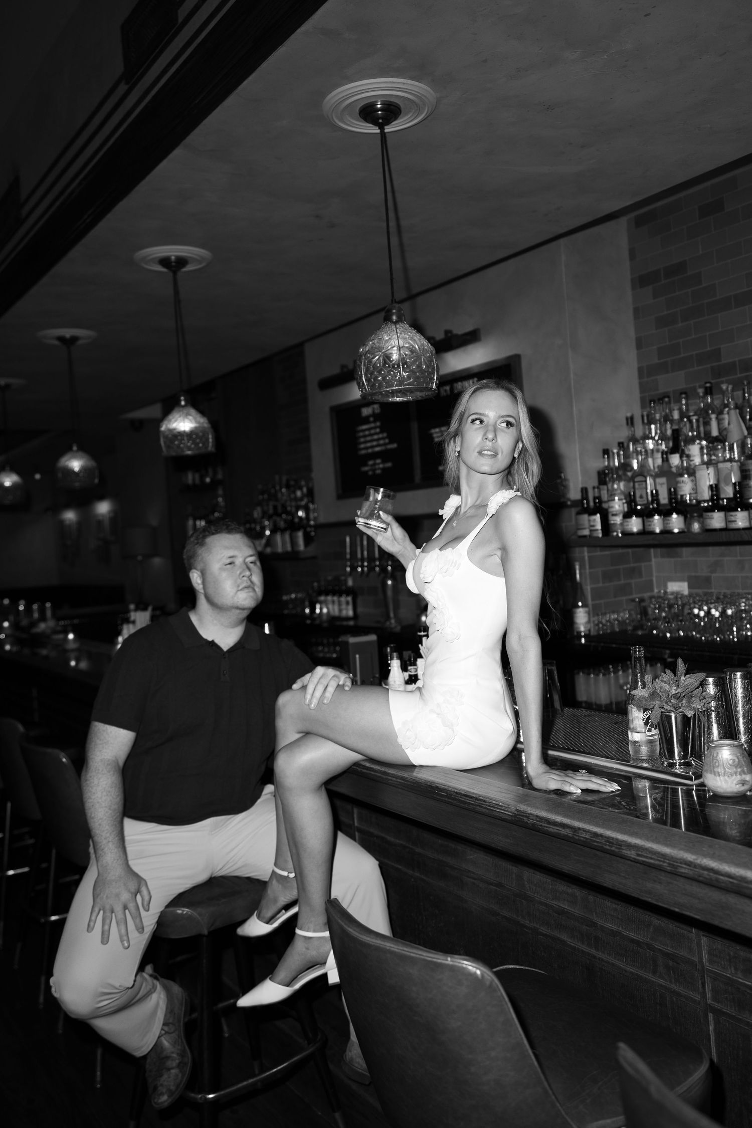 Black and white photo of a couple at a bar, woman in white dress holding a drink, man seated beside her.