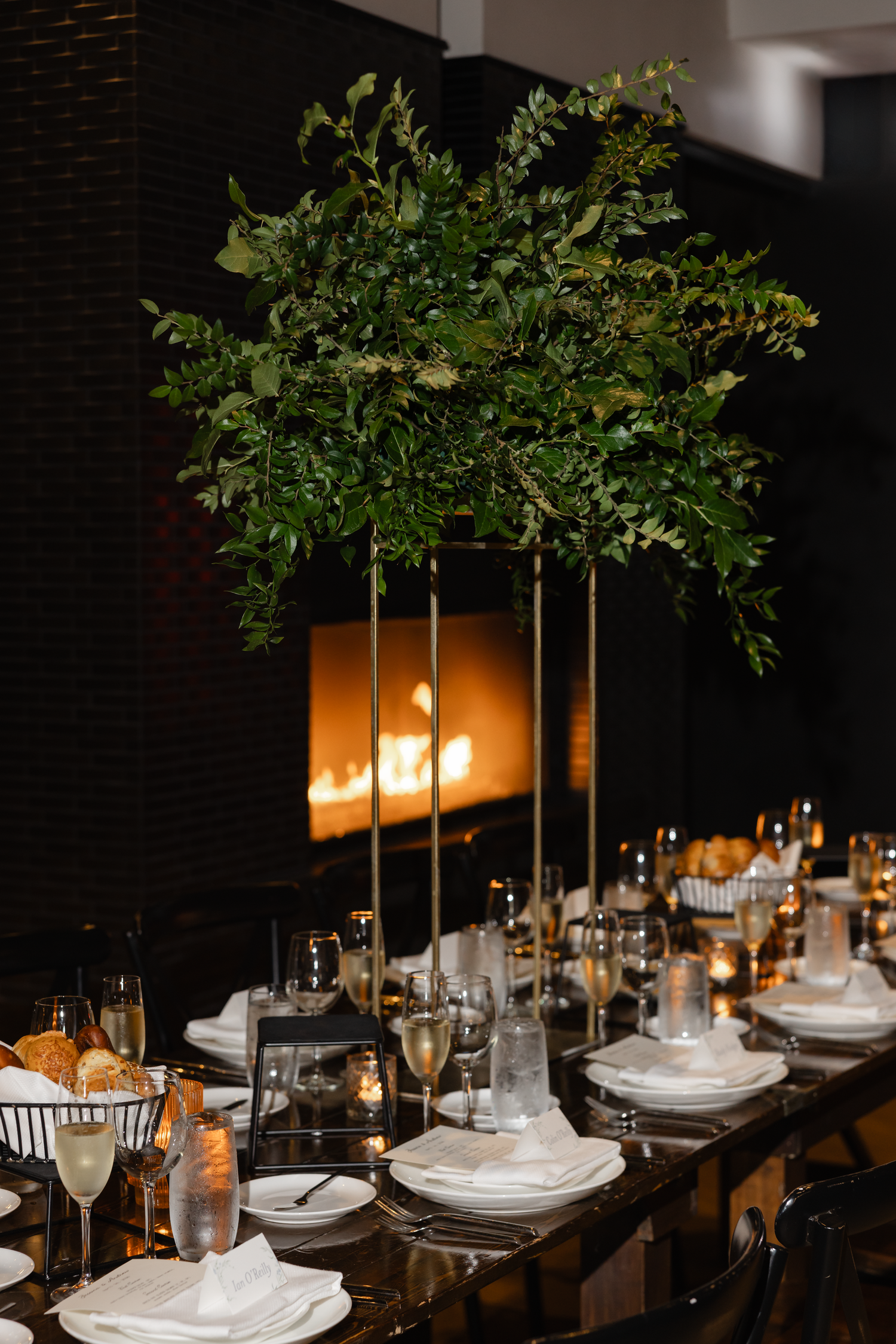 Elegant wedding reception table with tall greenery centerpiece, white plates, gold accents, and a glowing fireplace in the background.