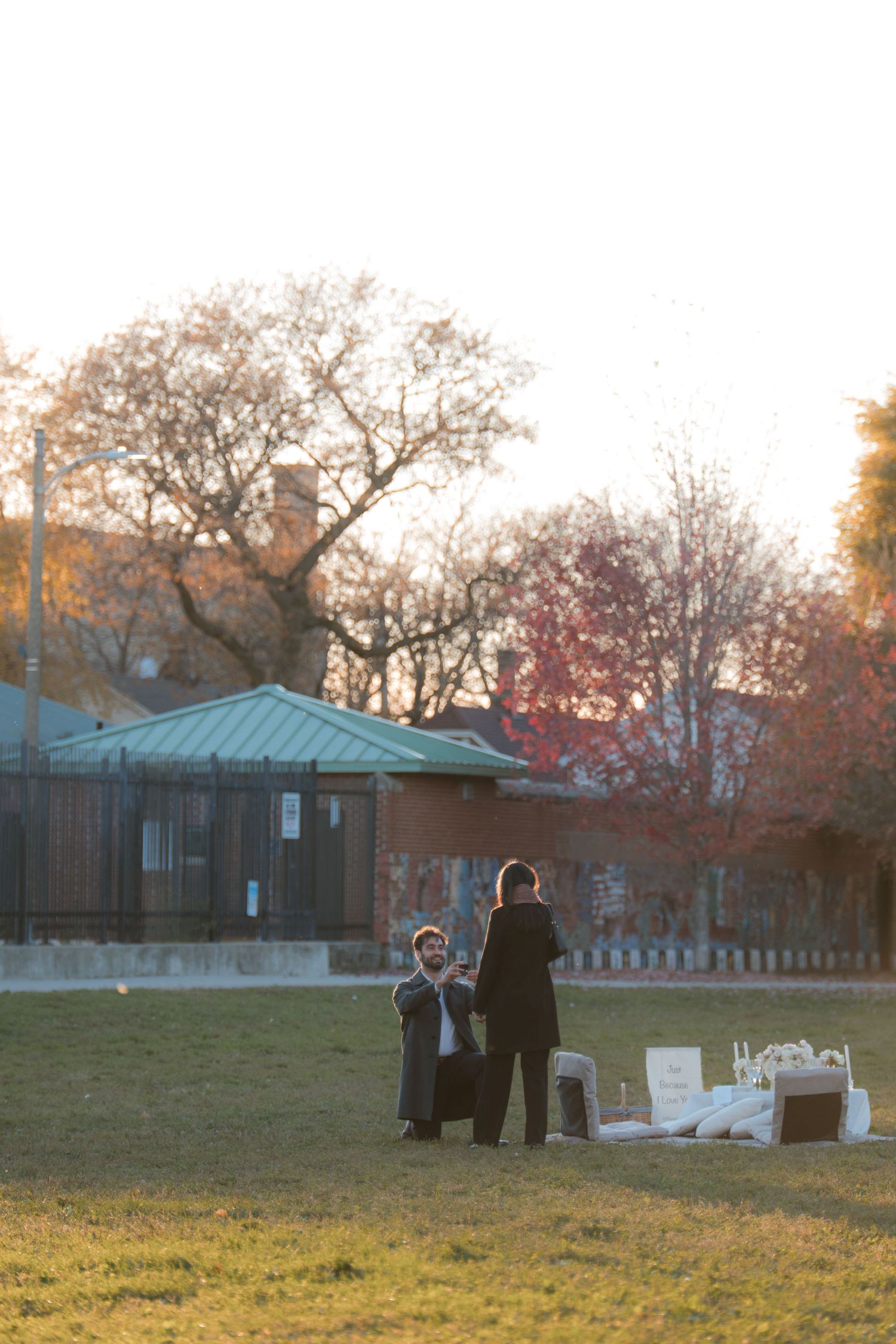 Two people standing on a grassy park at golden hour, with autumn trees and a brick building in the background.