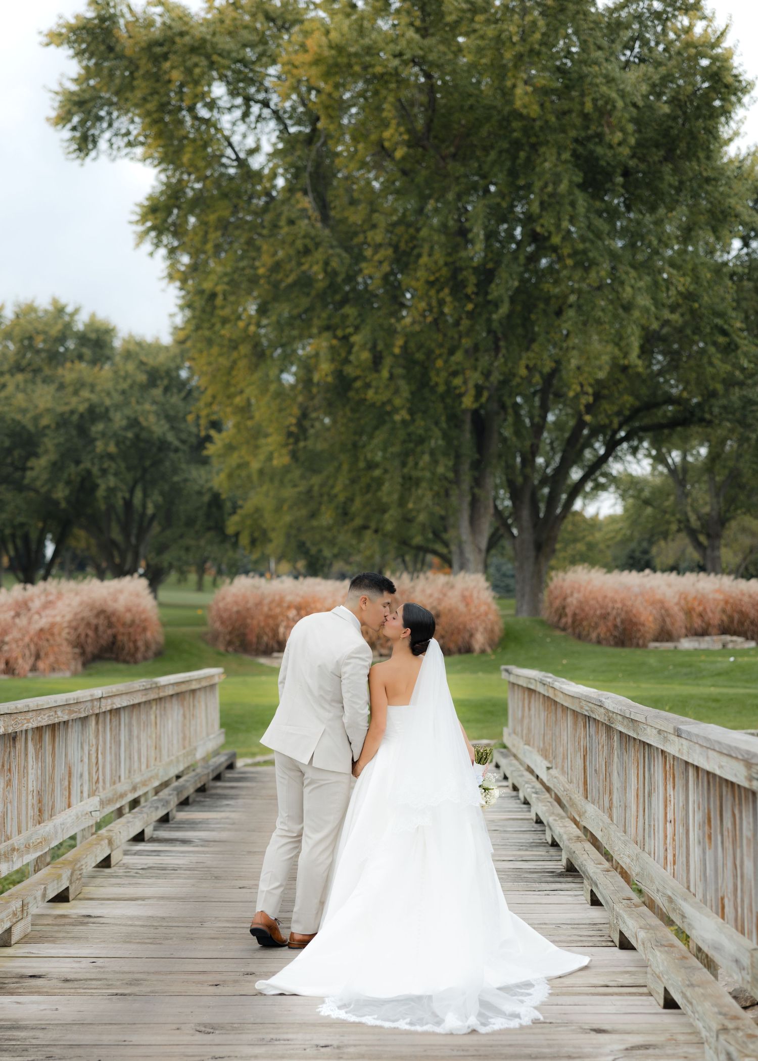 A wedding couple shares an intimate moment on a wooden bridge with trees and ornamental grasses.