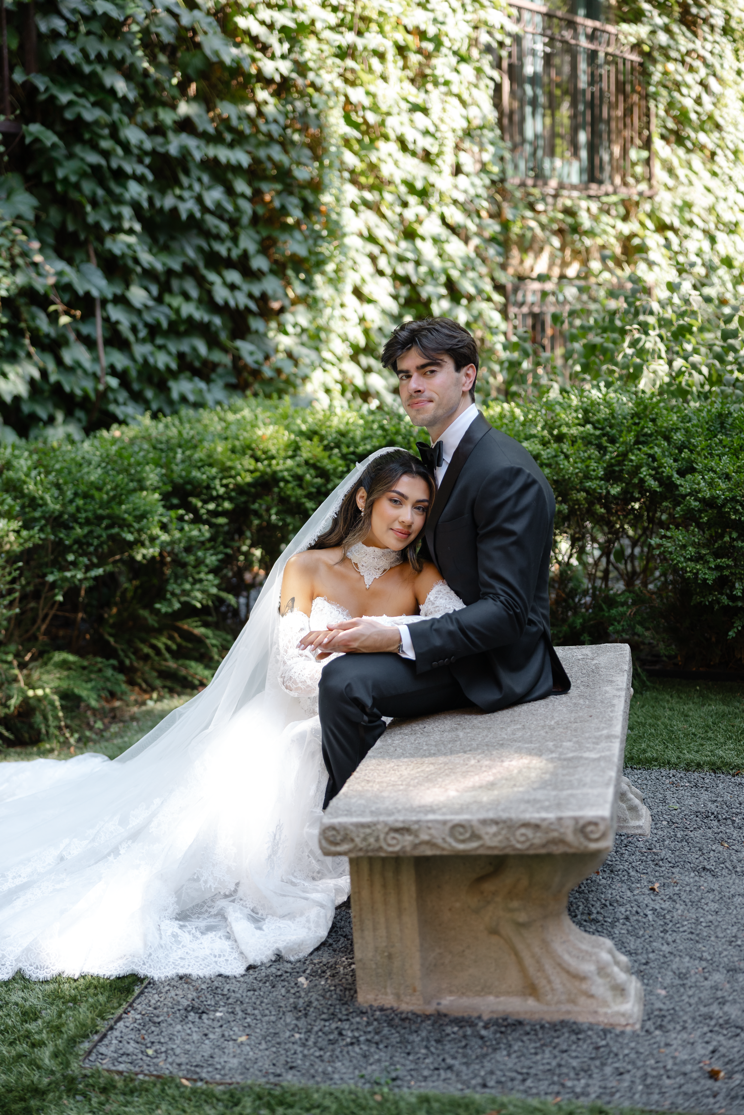 Bride and groom sit together on a stone bench in a lush garden, bride in white gown with veil, groom in dark suit.