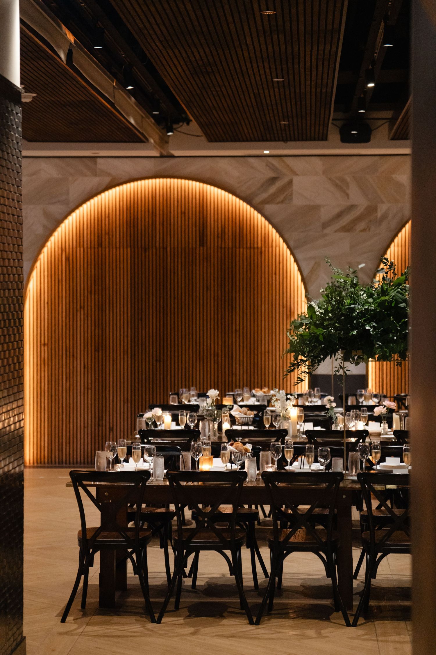 Elegant restaurant dining room with dark wood chairs, set tables, and a warm-lit arched wood panel feature wall.
