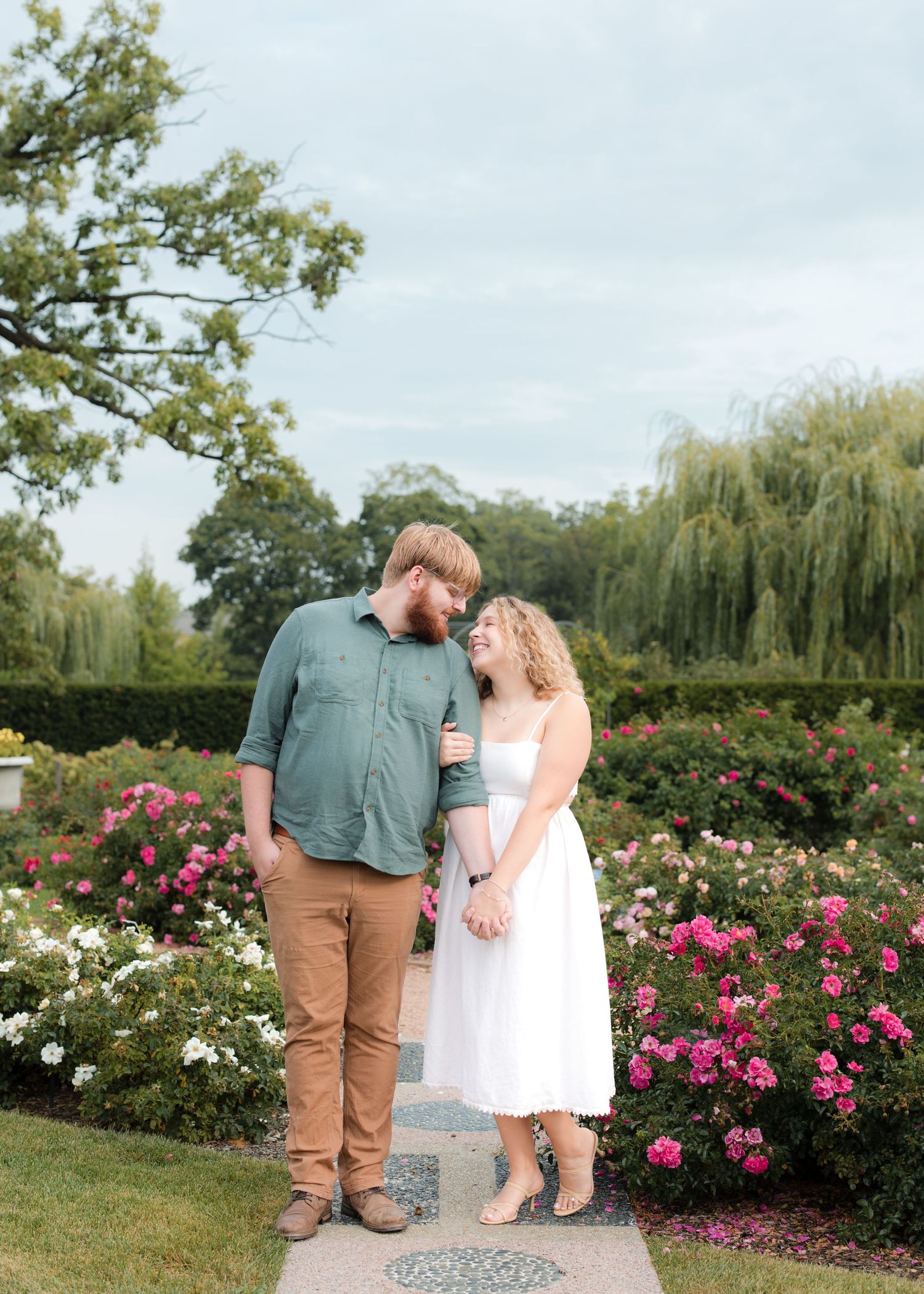 Couple laughing together in a lush garden surrounded by pink and white roses on a cloudy day.