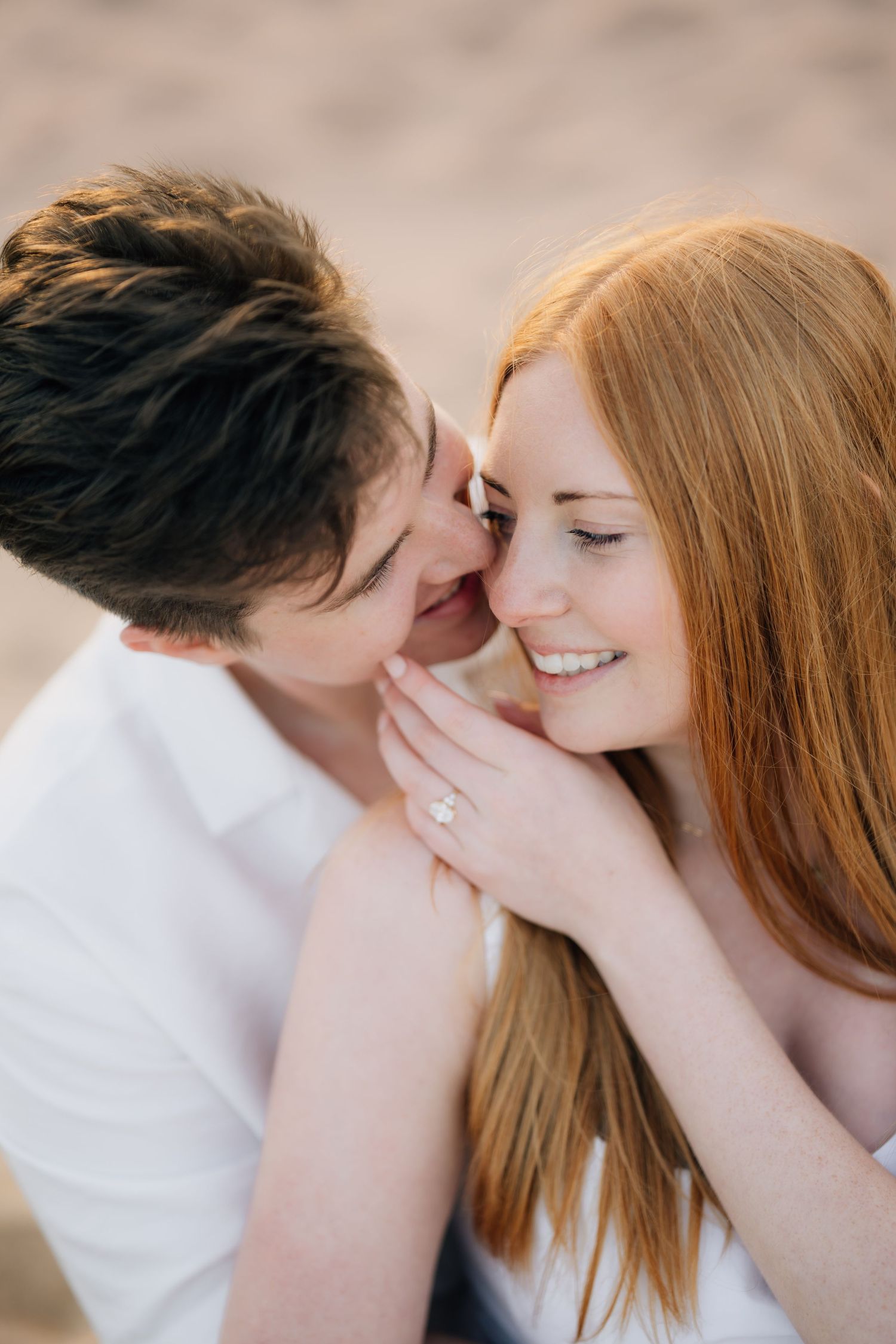 Couple sharing a tender moment on a beach, woman with red hair smiling and wearing an engagement ring.