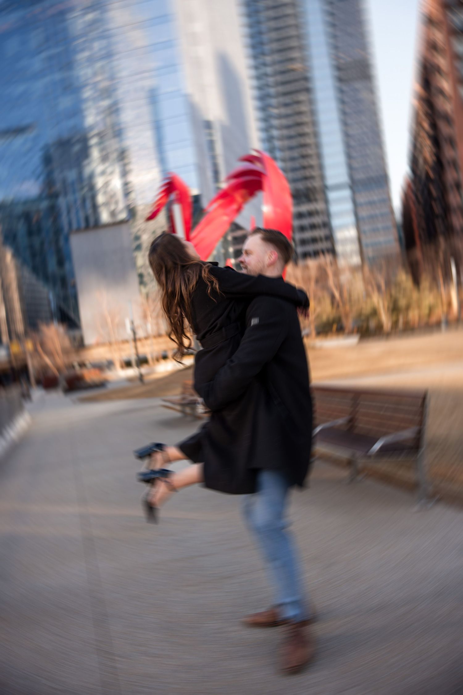 Couple embracing and spinning joyfully in an urban plaza with modern skyscrapers and a red sculpture backdrop.