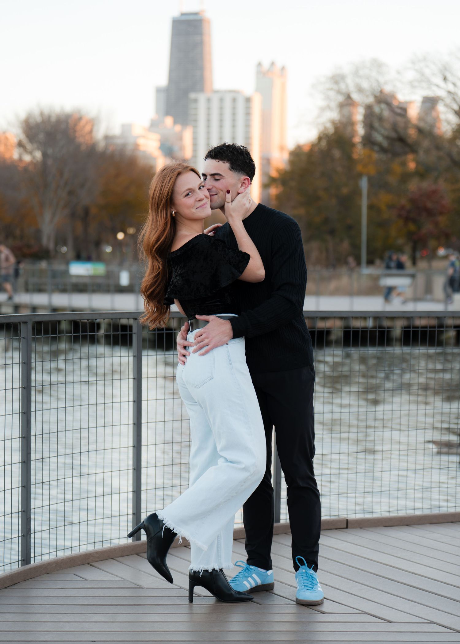 Couple embracing on waterfront boardwalk with Chicago skyline backdrop, man kissing woman's cheek in autumn setting.