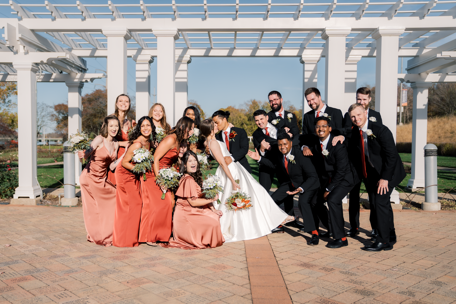 Wedding party lined up under white pergola wearing coral and black formal attire.
