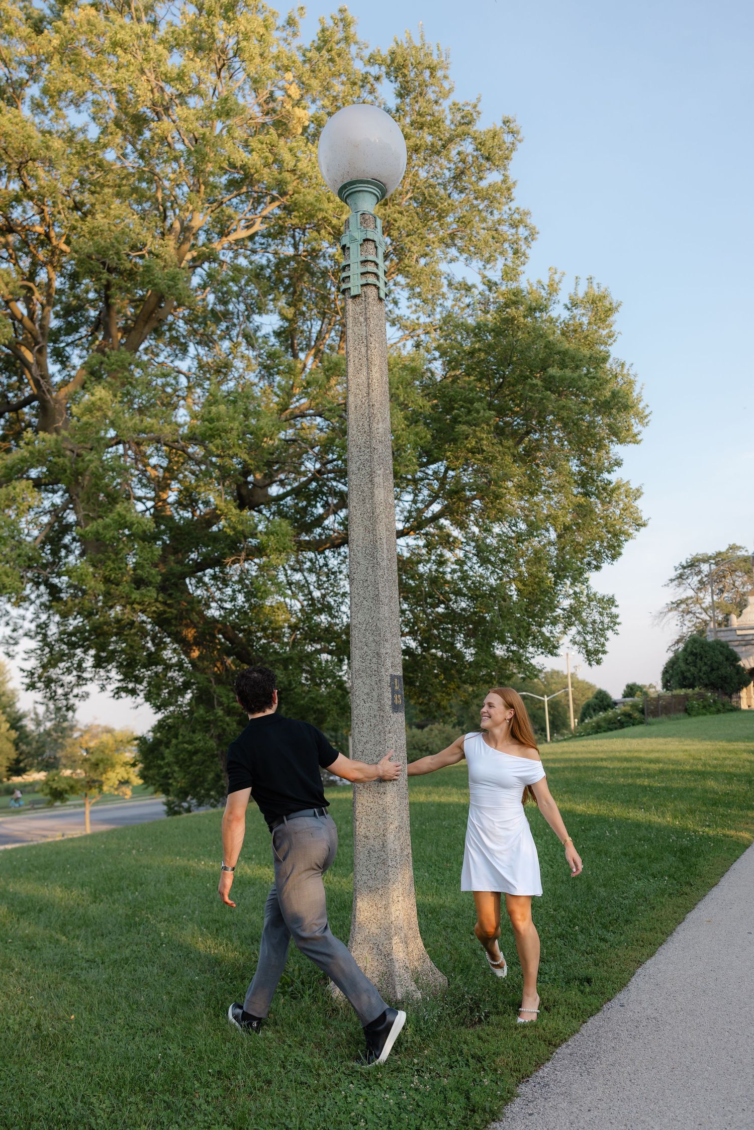 Couple playfully spinning around a vintage green lamppost on a grassy park path surrounded by lush trees.