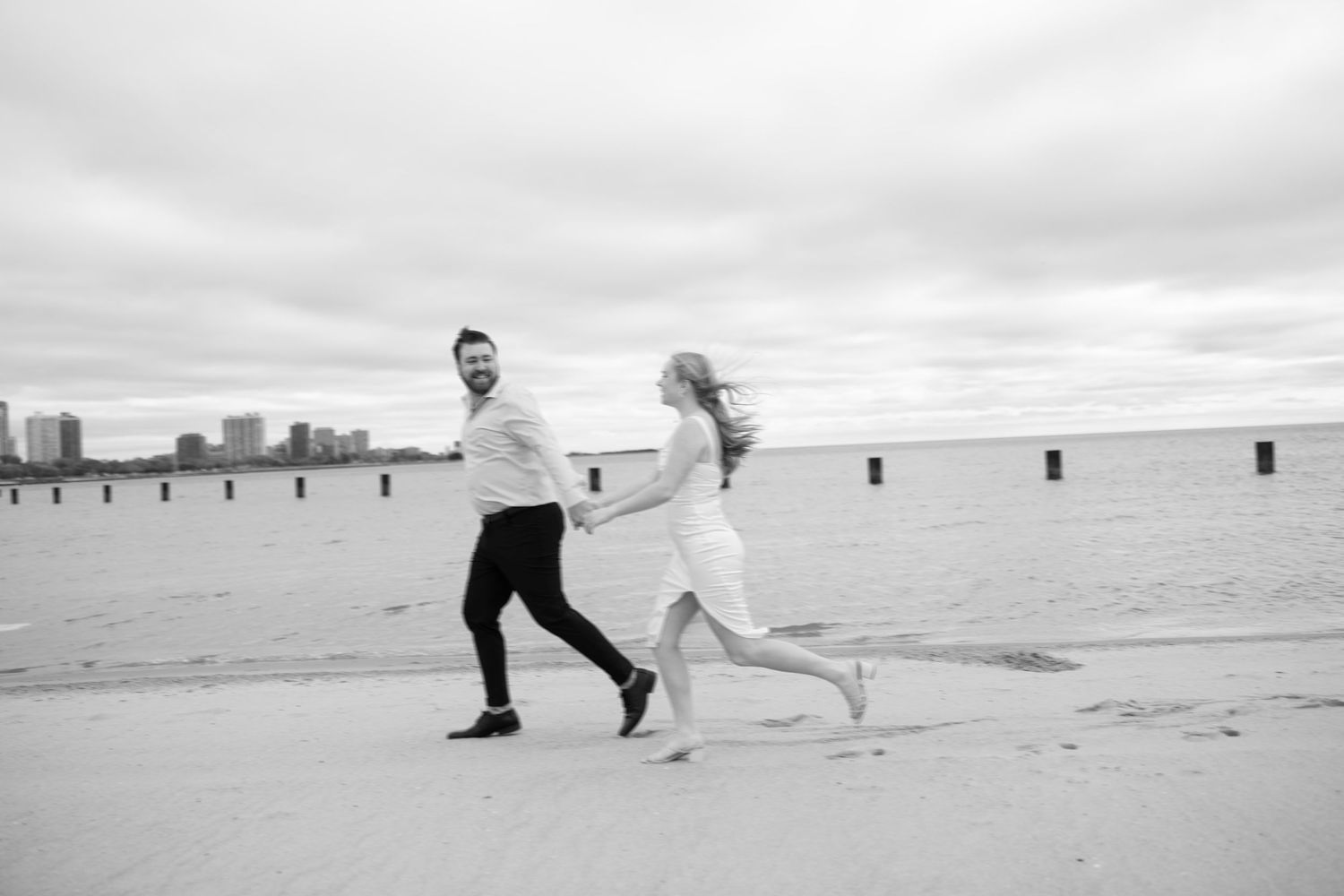 Black and white photo of a couple holding hands and running joyfully on a beach with a city skyline in the background.