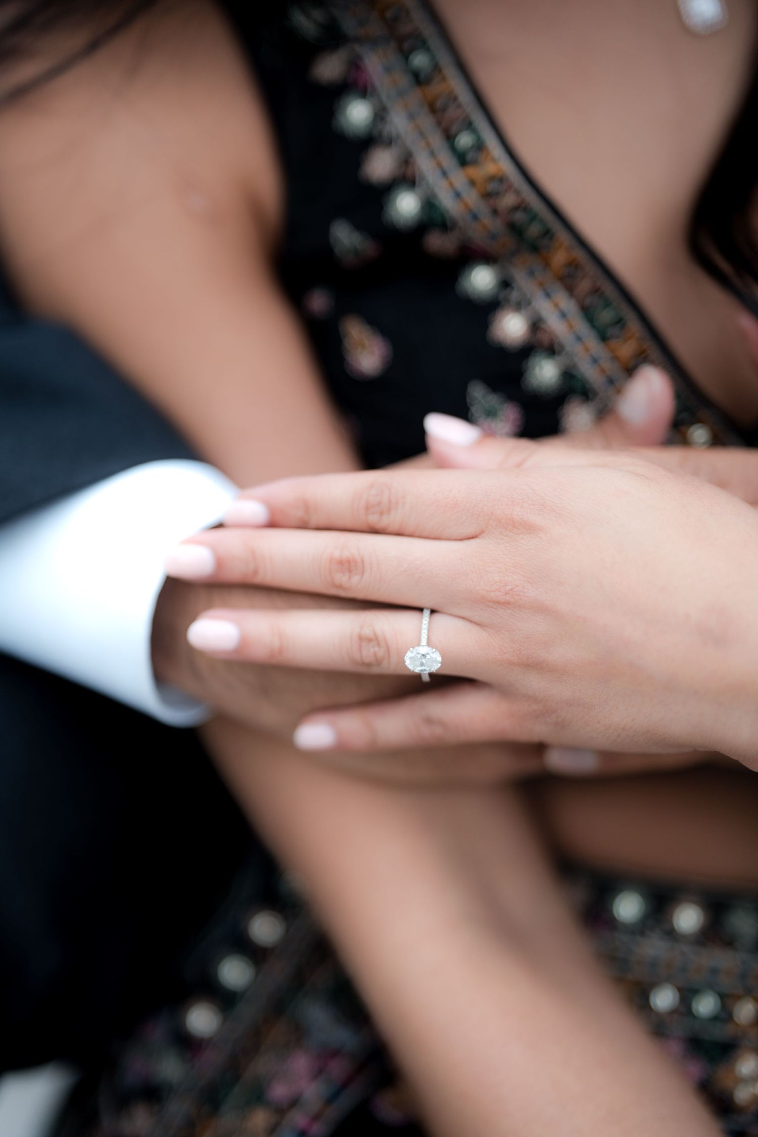 Close-up of a woman's hand wearing a stunning diamond engagement ring, resting on a man's arm in a white suit.