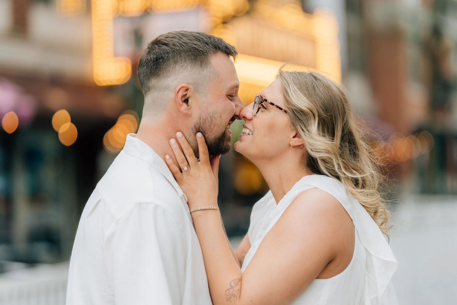 Couple sharing a romantic almost-kiss outdoors, woman in white dress with glasses, man in white shirt, bokeh city lights background.