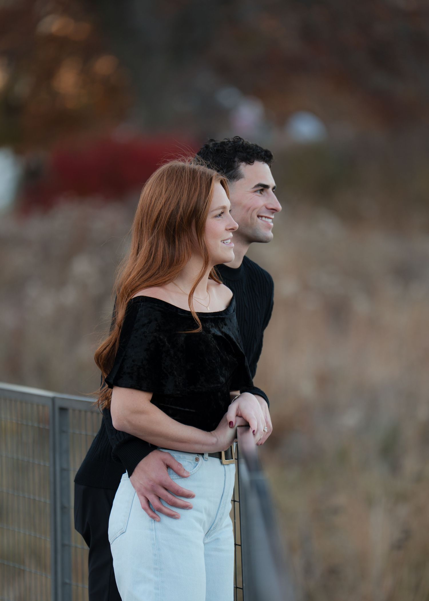 A smiling couple embraces outdoors near a metal railing, surrounded by autumn foliage in warm fall colors.