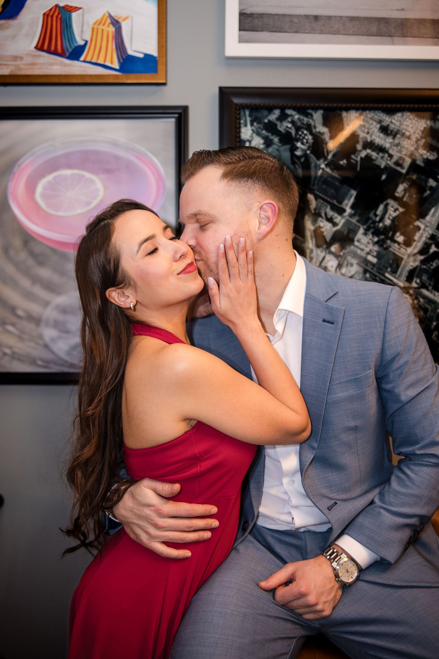 A couple shares a romantic kiss in an art gallery, woman in red dress and man in gray suit surrounded by colorful paintings.