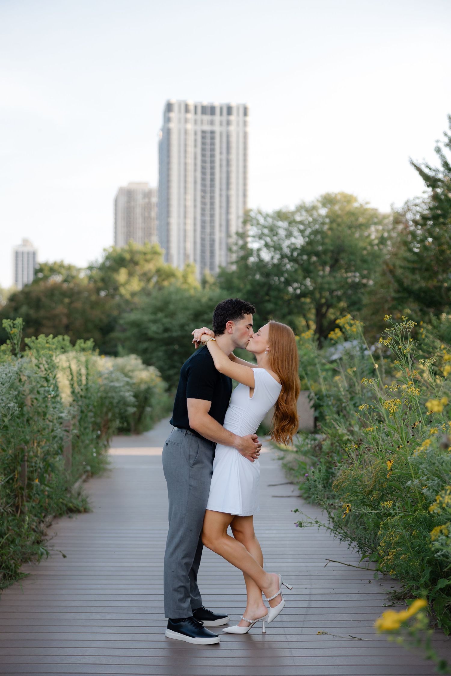 Couple sharing a romantic kiss on a wooden boardwalk path surrounded by lush greenery with city skyline backdrop.