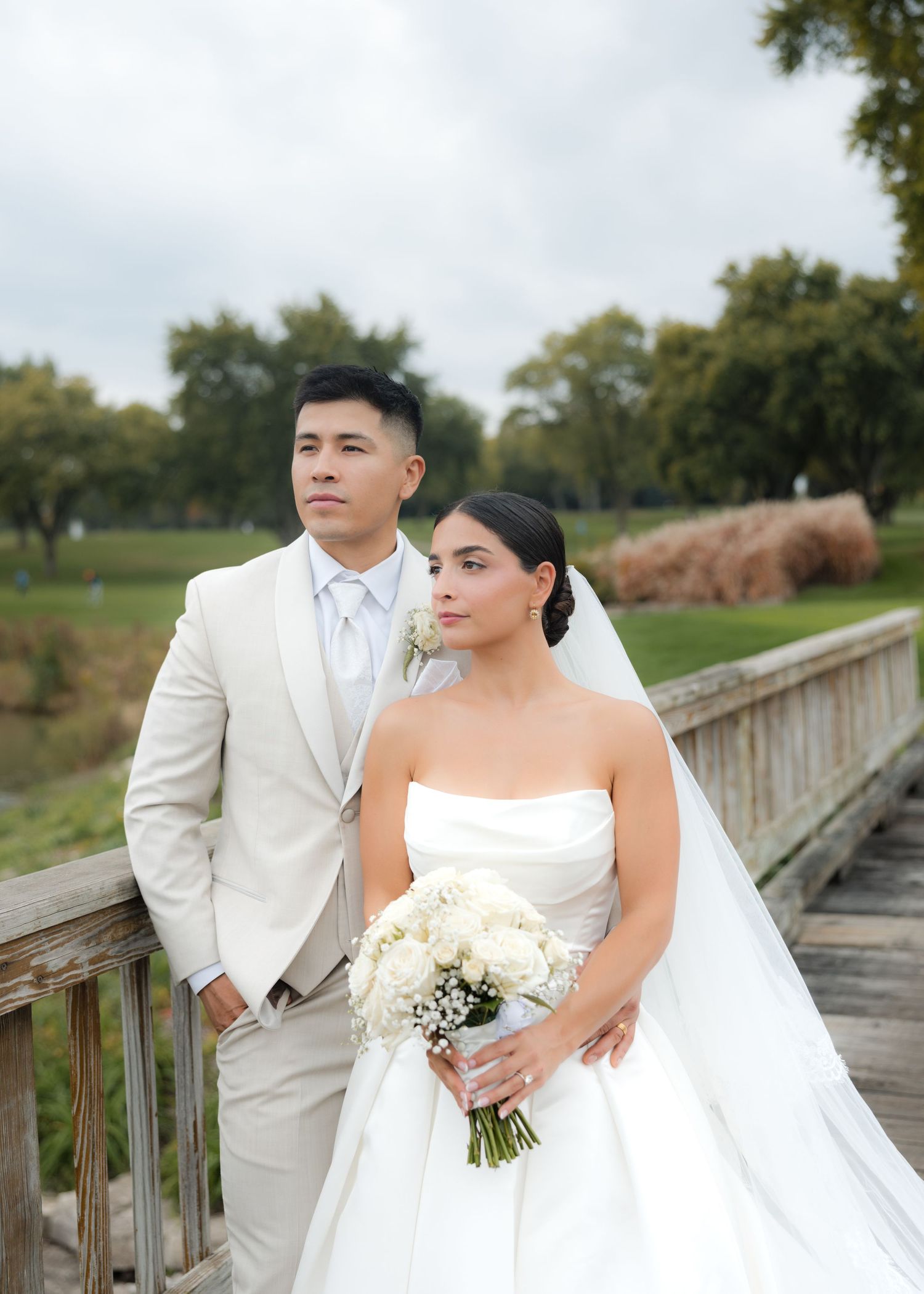 A wedding couple poses on a wooden bridge surrounded by autumn foliage in an outdoor park setting.