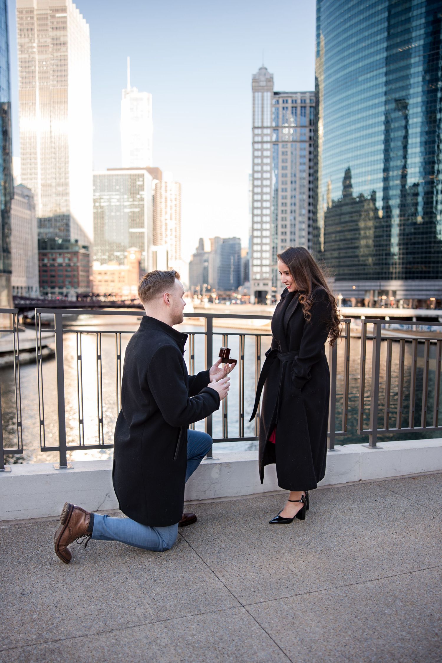 Man proposing on one knee with ring box to woman on urban riverwalk with city skyline backdrop.