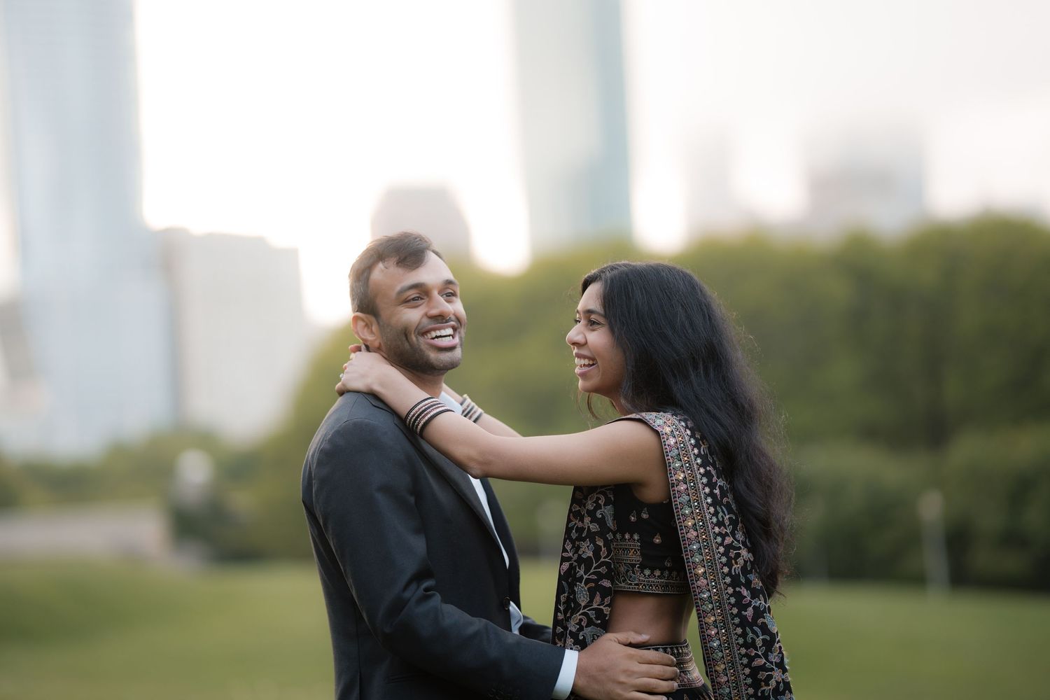 Happy South Asian couple embracing outdoors in formal attire with city skyline in background.