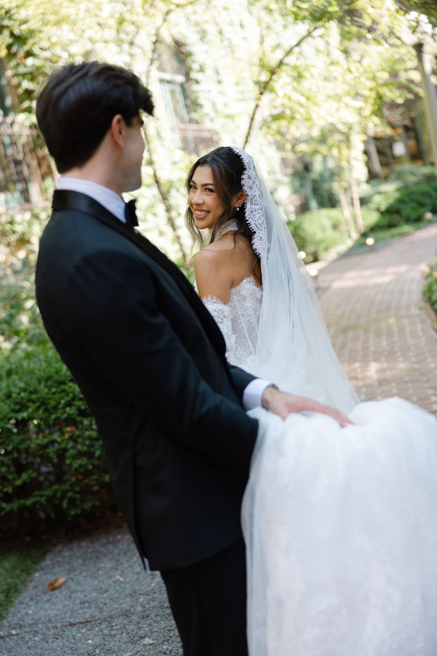 Smiling bride in lace gown and veil holds groom's hand in a sunlit garden during outdoor wedding photos.