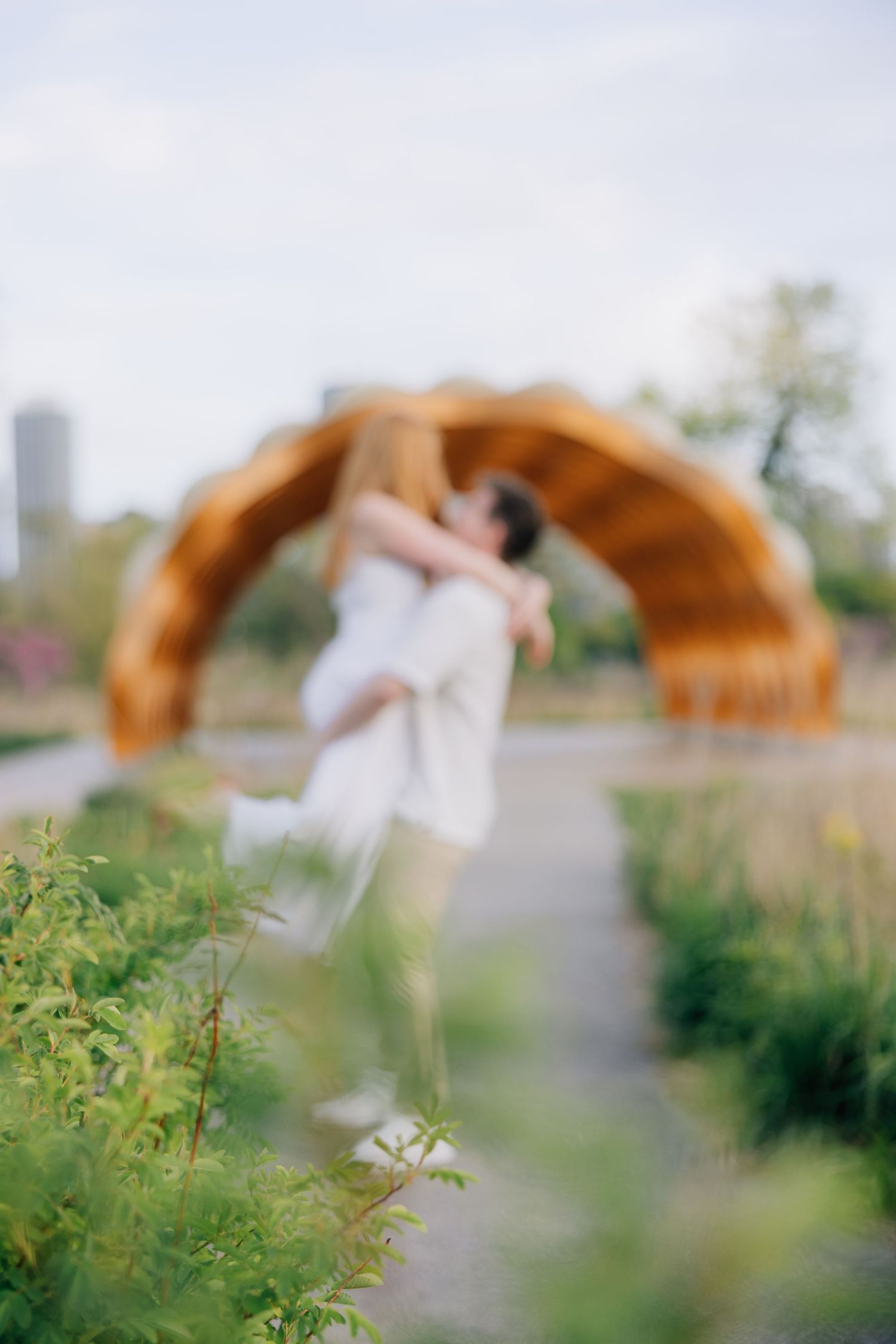 Couple embracing in a lush garden path with a wooden arch structure in the background on a cloudy day.
