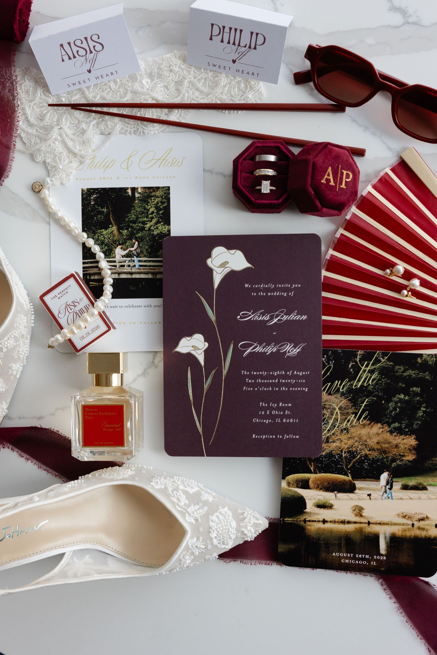 Elegant wedding flat lay with burgundy invitations, white heels, perfume, pearl necklace, rings, red fan, and sunglasses.