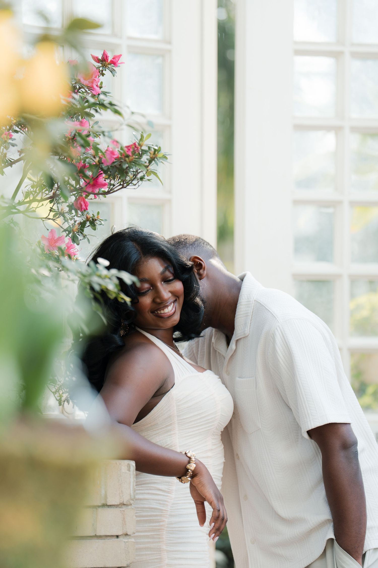 A joyful Black couple in matching white outfits share a tender moment surrounded by pink flowers near bright windows.